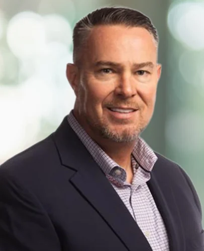 A man with short gray hair and a trimmed beard, wearing a dark blazer over a checked shirt, poses in front of a blurred light background.