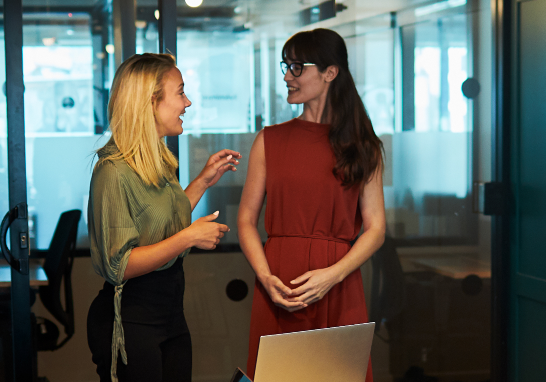 Two women stand and talk in a modern office, one wearing a green blouse and black pants, the other in a sleeveless red dress. A laptop sits open on a table nearby.