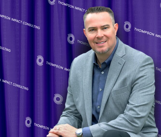 Man in a gray suit jacket sits in front of a purple backdrop with "THOMPSON IMPACT CONSULTING" and circular logos printed repeatedly.