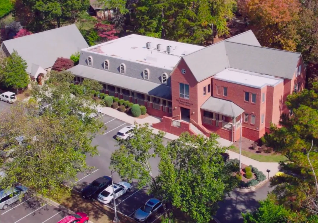 Aerial view of a two-story red brick building with white trim, surrounded by trees and a parking lot with several cars.