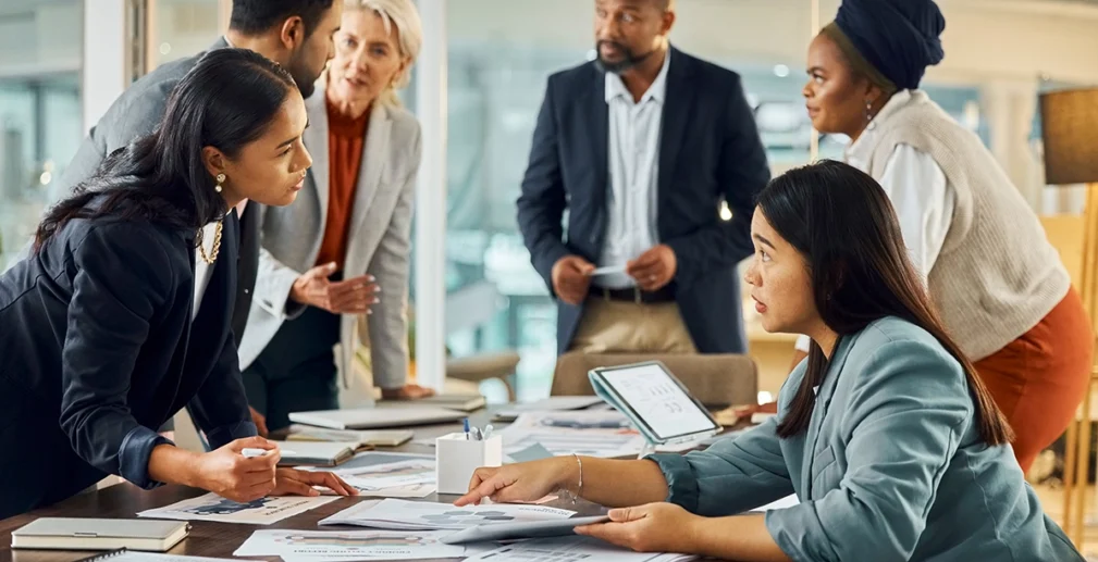 Six people in business attire have a discussion around a table covered with documents and charts in a modern office setting.