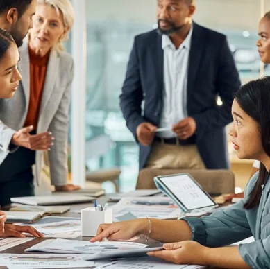 Six people in business attire have a discussion around a table covered with documents and charts in a modern office setting.