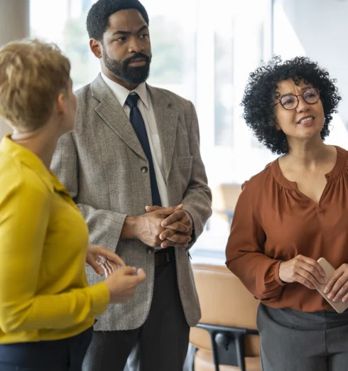 Three professionally dressed people are standing indoors, engaged in a conversation.