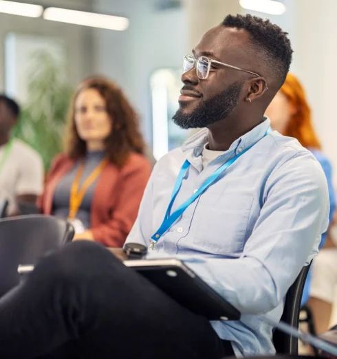 Man with glasses and a blue lanyard sits in a chair holding a tablet, attending what appears to be a seminar or conference with other people in the background.