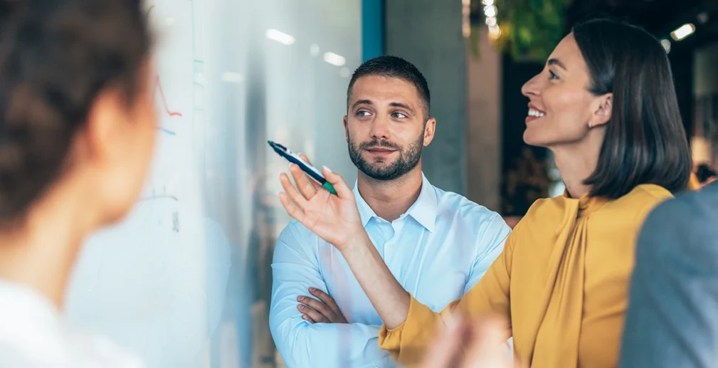 A woman points at a whiteboard while discussing with two colleagues; one man looks on with arms crossed, and another person is partially visible in the foreground.
