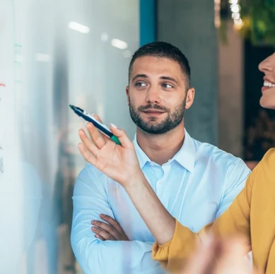 A woman points at a whiteboard while discussing with two colleagues; one man looks on with arms crossed, and another person is partially visible in the foreground.