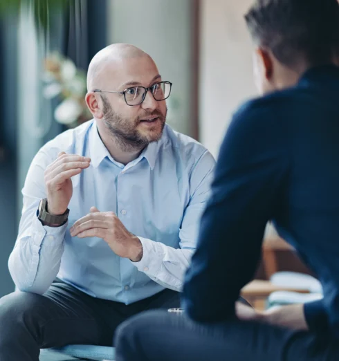 Two men sit indoors having a conversation. One man, wearing glasses and a light blue shirt, gestures with his hands while speaking to the other man, who has his back to the camera.