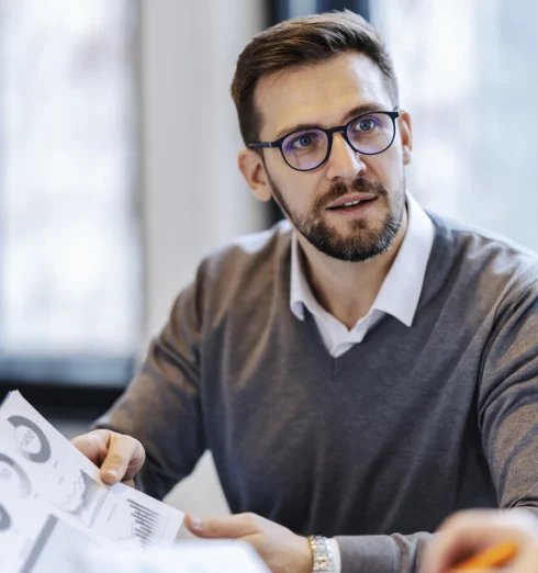 Man with glasses and beard holding documents, looking attentively at someone during a discussion in an office setting.