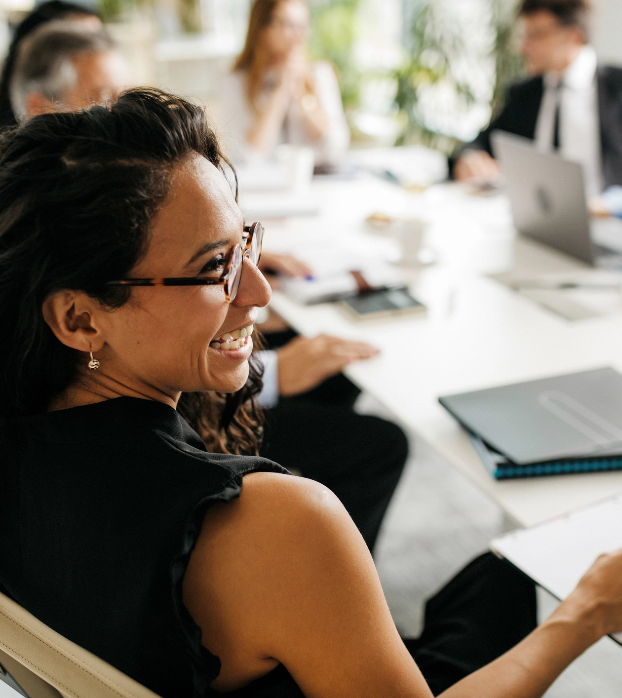 A woman wearing glasses smiles during a business meeting at a conference table with laptops, notebooks, and other people in the background.