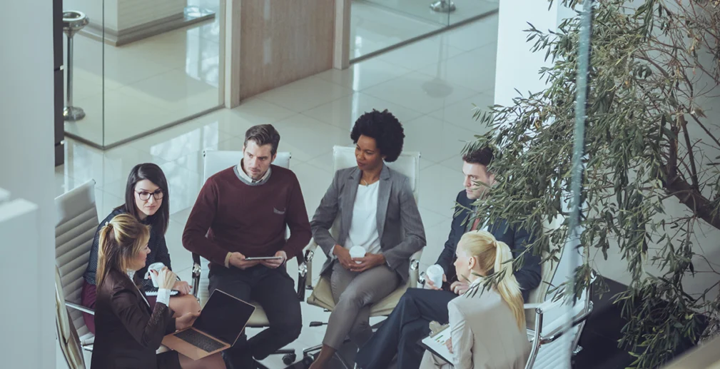 Six people in business attire sit in a circle having a discussion in a modern office with glass walls and natural light.