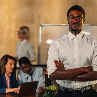 A man stands with arms crossed in the foreground, while two pairs of colleagues have discussions and work on laptops in an office setting with a whiteboard in the background.