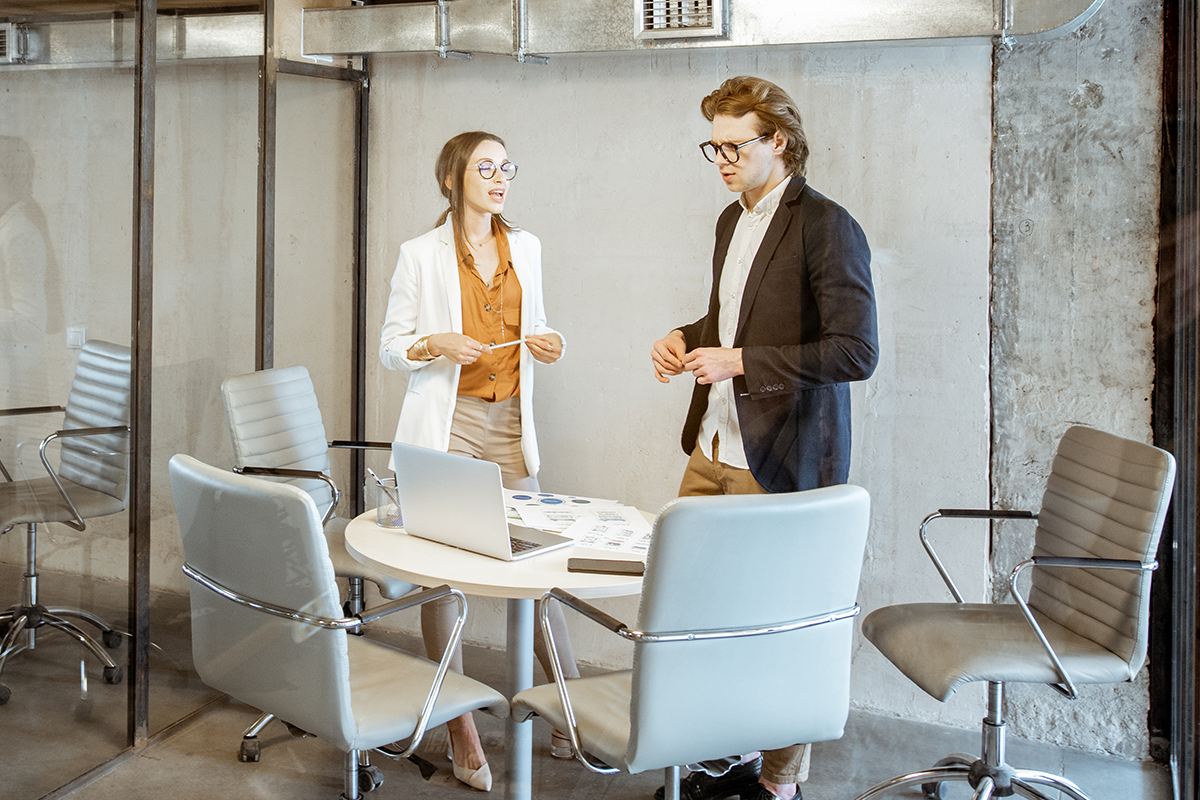 Two people stand by a small round table with a laptop and documents in a modern office meeting room with gray chairs.