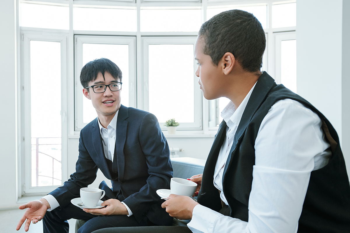 Two people in business attire sit and talk while holding coffee cups in a bright room with large windows.