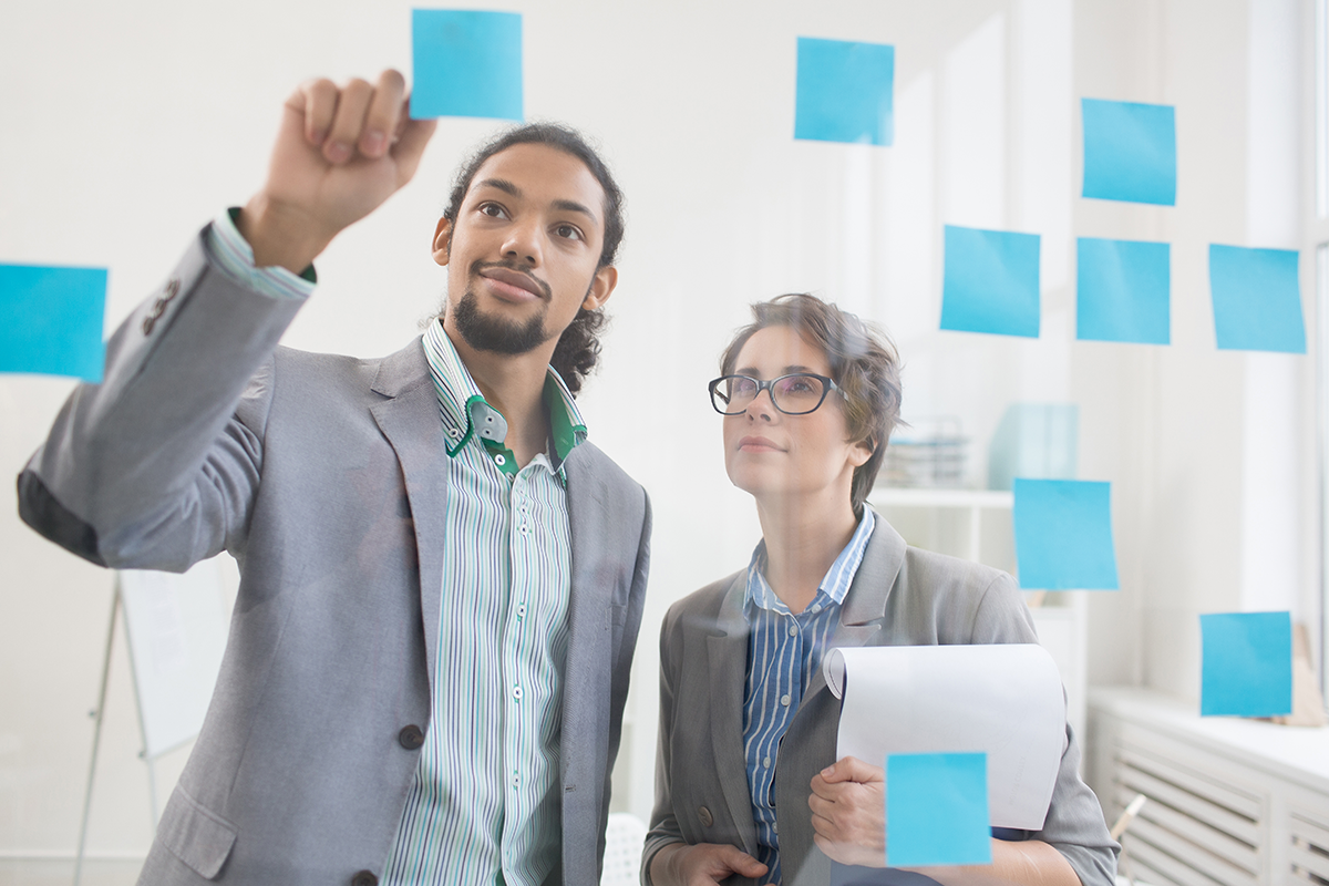 Two people in business attire look at blue sticky notes on a glass wall. One person is placing a note, while the other holds documents and observes. Office setting with natural light.