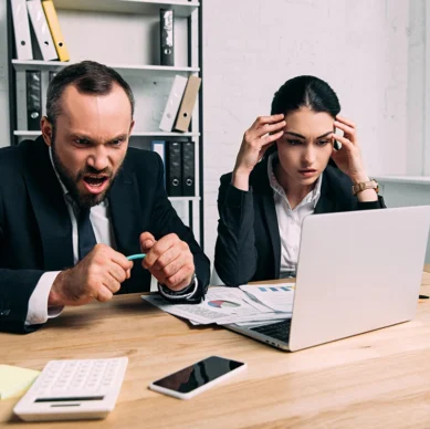 Two business professionals sitting at a desk, looking frustrated while staring at a laptop screen with documents, calculator, and phone on the table.