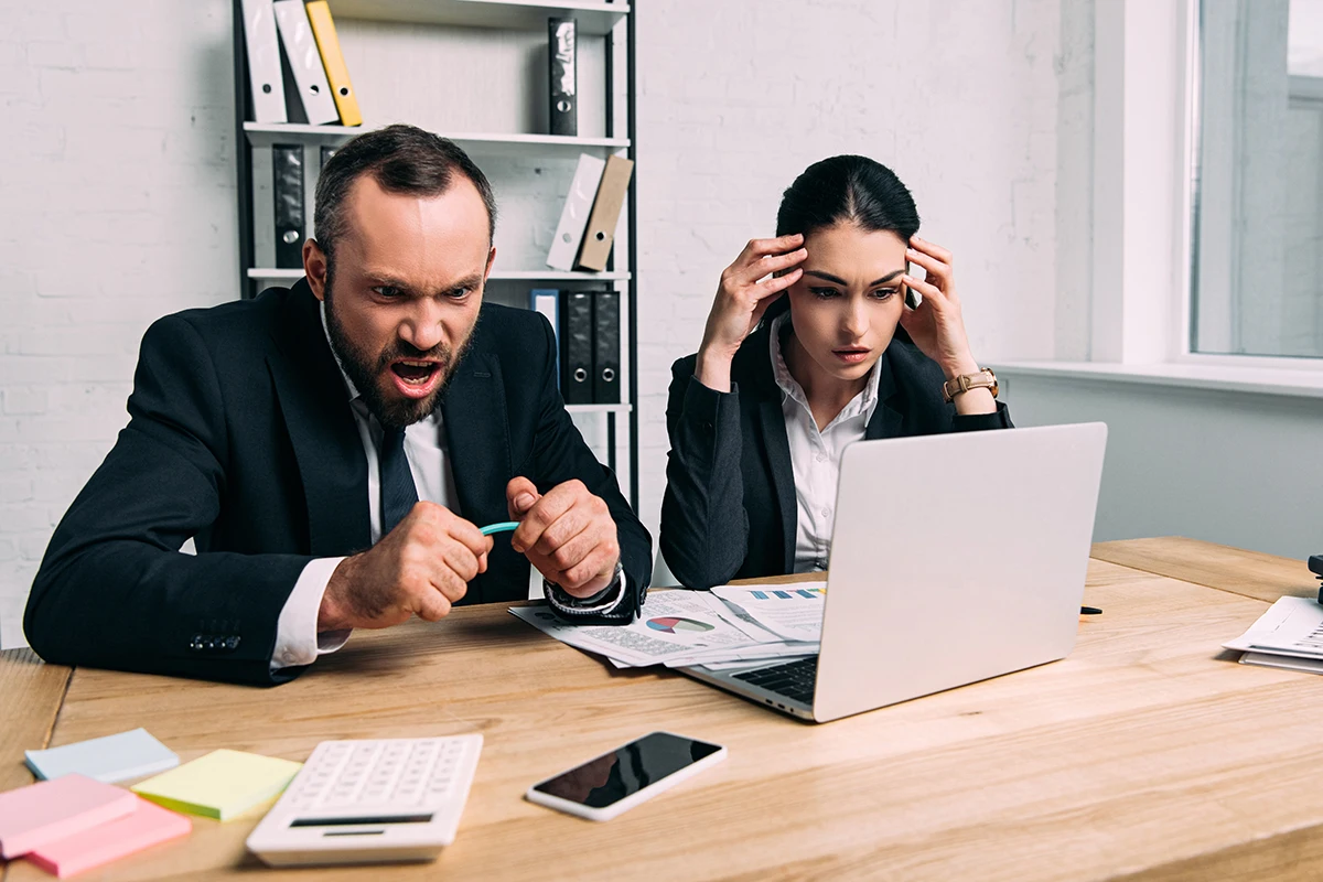 Two business professionals sitting at a desk, looking frustrated while staring at a laptop screen with documents, calculator, and phone on the table.