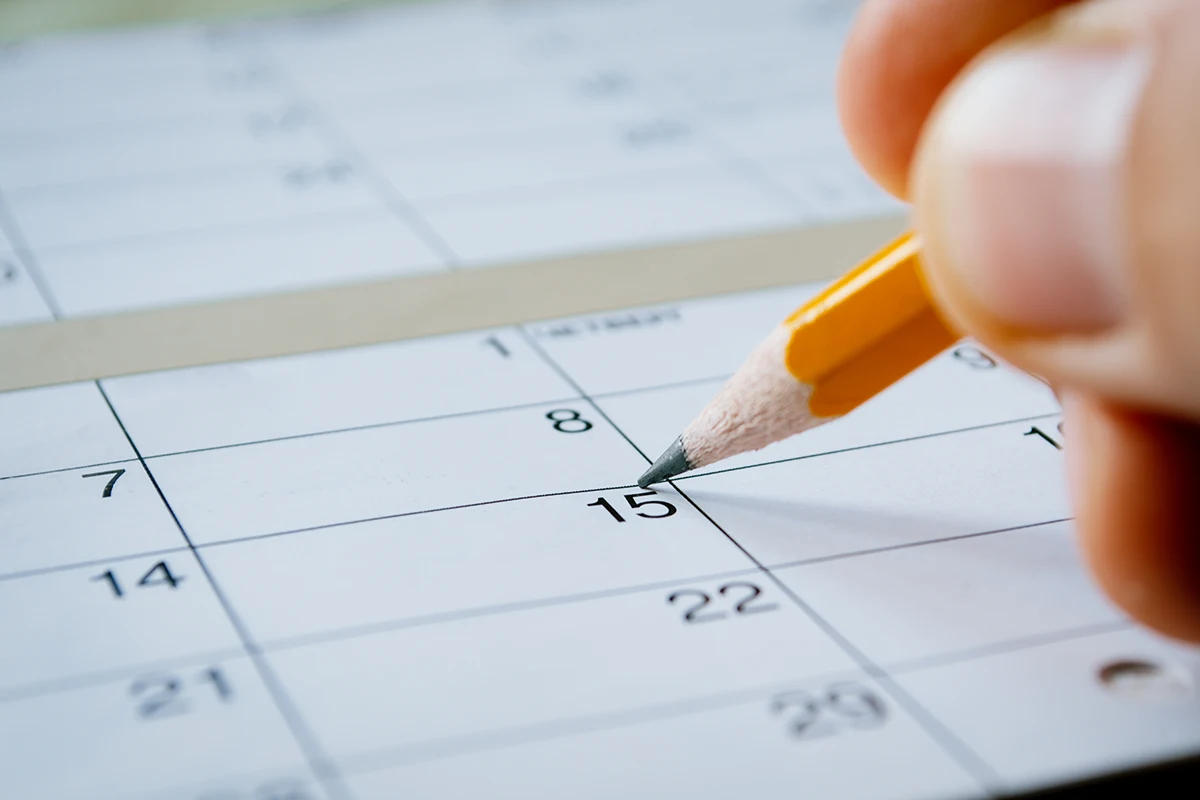 A hand holding a yellow pencil points to the 15th day on a paper calendar, with part of the month and dates visible in the background.