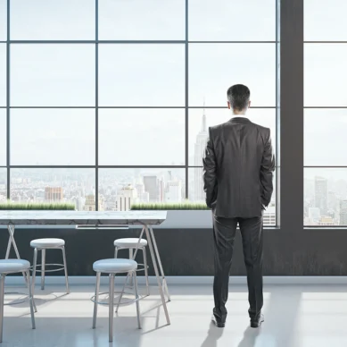 A man in a suit stands in a modern office with large windows, looking out at a city skyline. Four white chairs and a table are in the foreground.