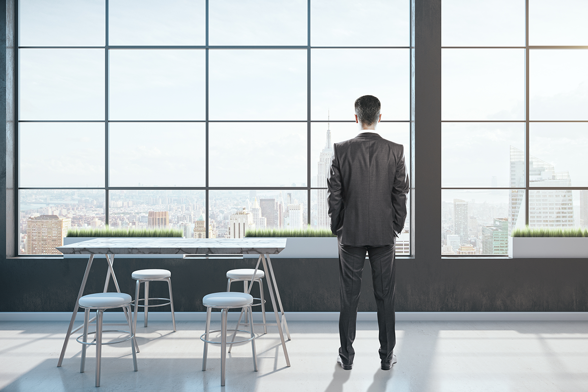 A man in a suit stands in a modern office with large windows, looking out at a city skyline. Four white chairs and a table are in the foreground.