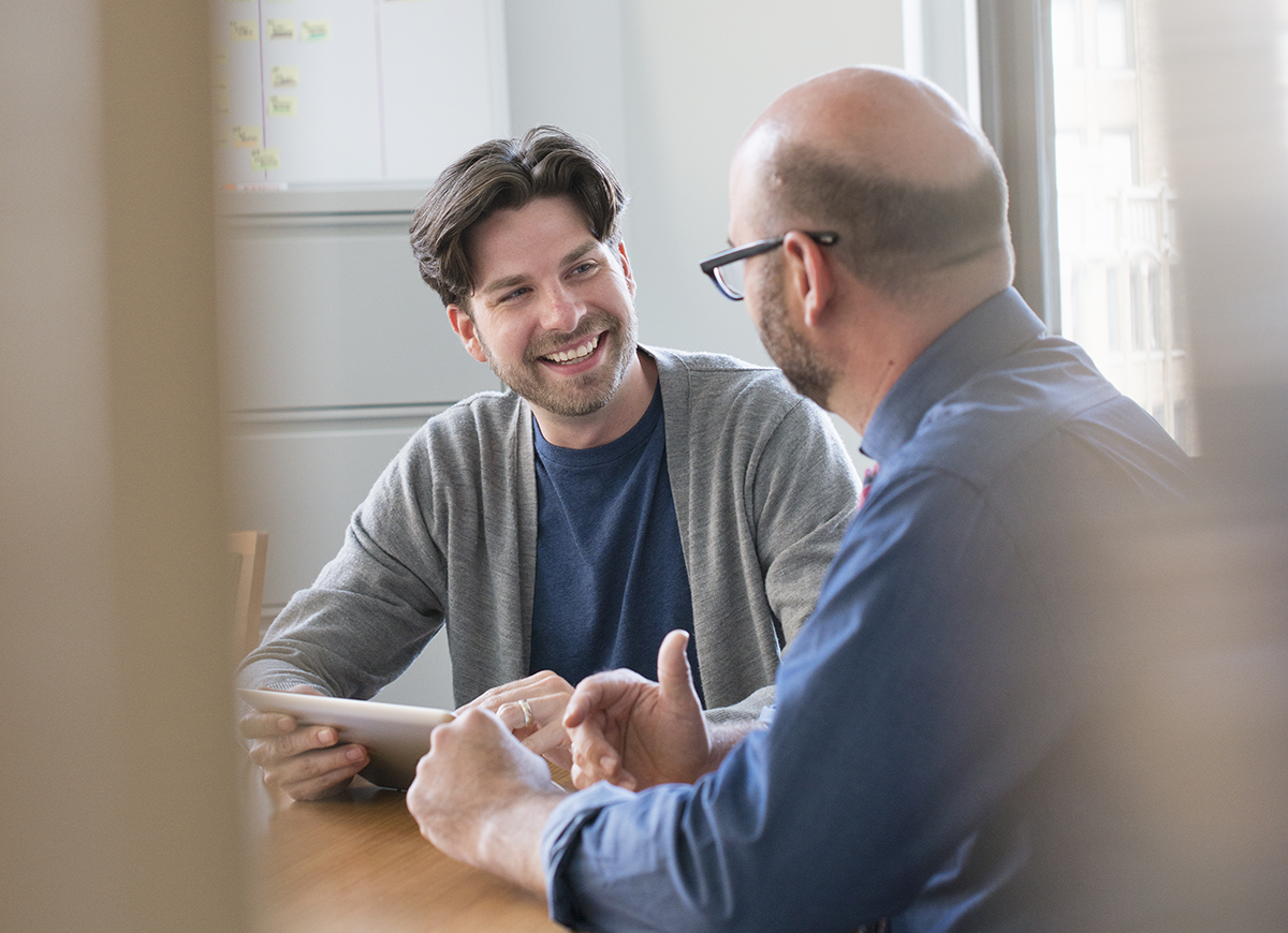 Two men sit at a table having a discussion. One is smiling and holding a tablet, while the other gestures with his hand. An office setting is visible in the background.