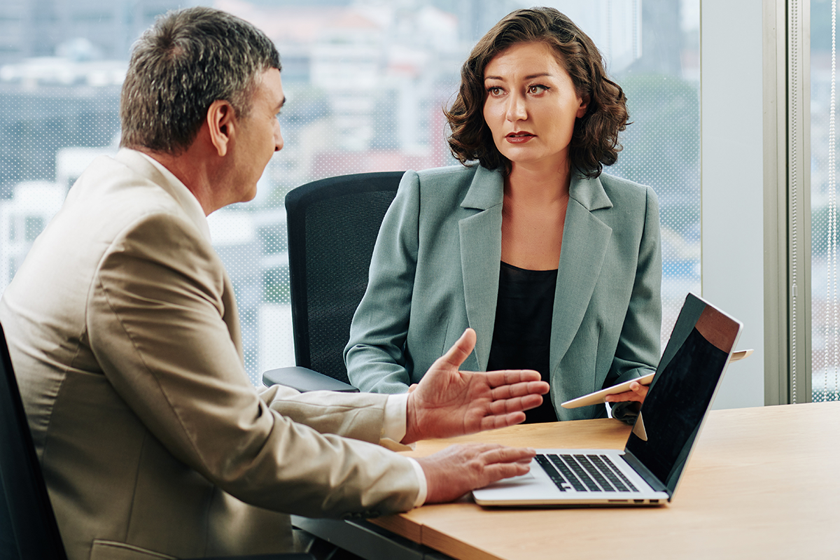 A man and a woman in business attire sitting across from each other at a desk with a laptop.
