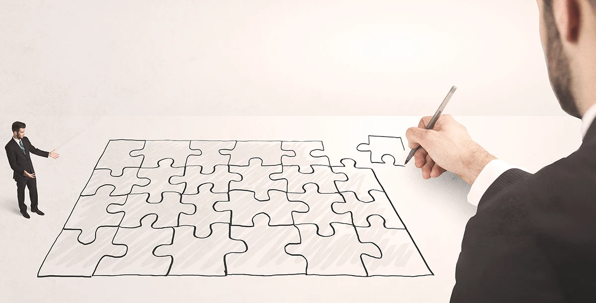 A man’s hand draws a puzzle piece onto a large puzzle, while a miniature man in a suit stands next to it.