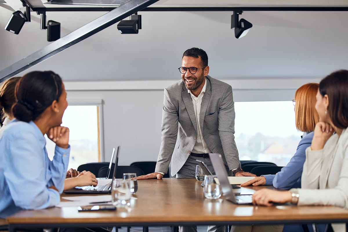 A man in a grey suit leads a meeting with four colleagues around a wooden conference table.
