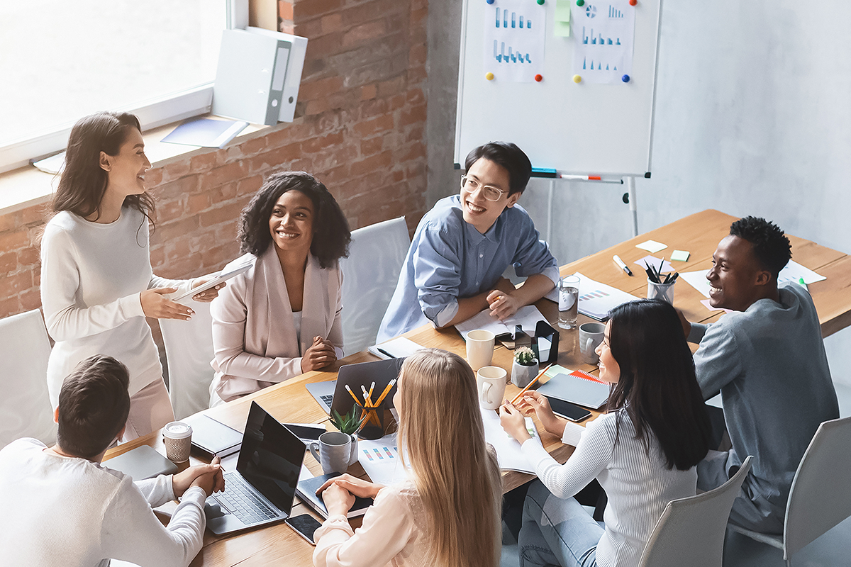 A diverse group of colleagues seated around a conference table listening to a woman standing and presenting.