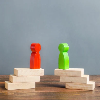 A red wooden figure and a green wooden figure stand on separate stacks of wooden blocks, facing each other, flanked by uncolored wooden figures.
