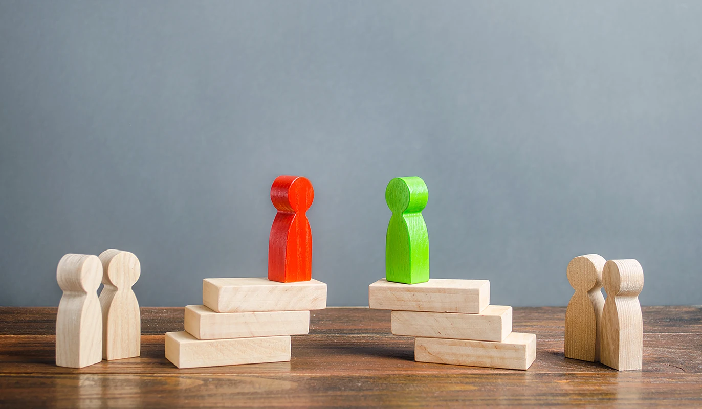 A red wooden figure and a green wooden figure stand on separate stacks of wooden blocks, facing each other, flanked by uncolored wooden figures.
